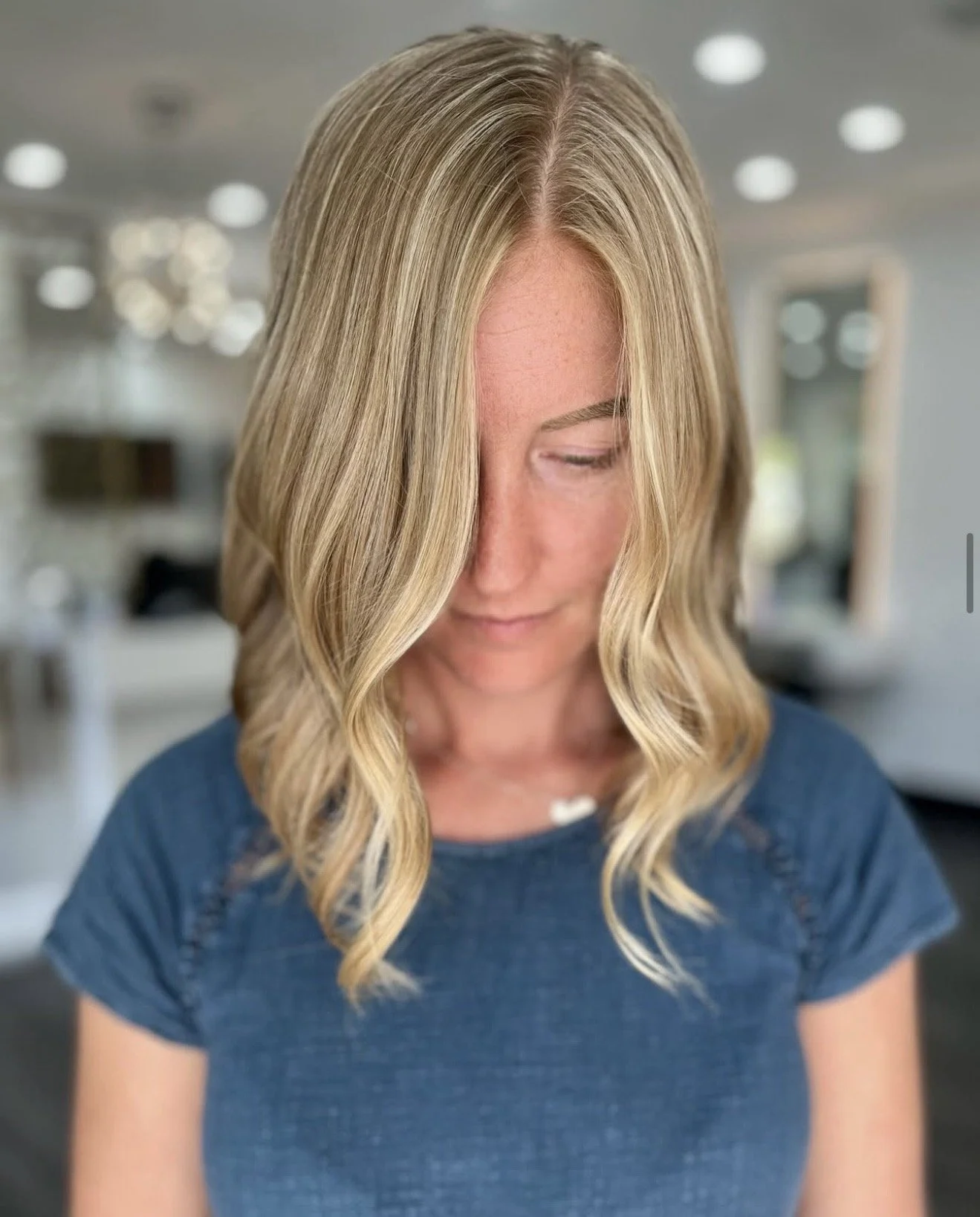 Person with shoulder-length wavy blonde hair in a blue shirt, looking down in a salon setting.