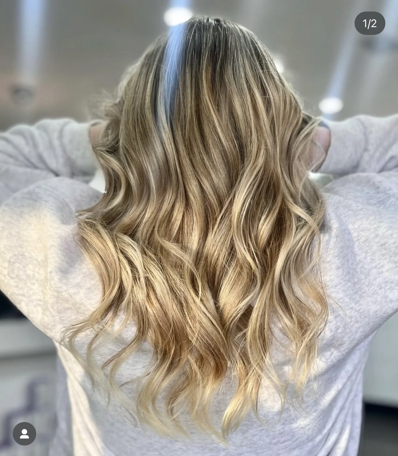 Woman with wavy blond hair from the back wearing a light sweater, arms raised, indoors.