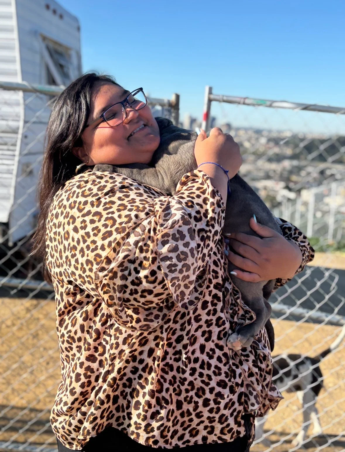 A woman with long dark hair and glasses, wearing a leopard print jacket, embraces and hugs a small black and gray puppy on an outdoor rooftop with a city skyline in the background.