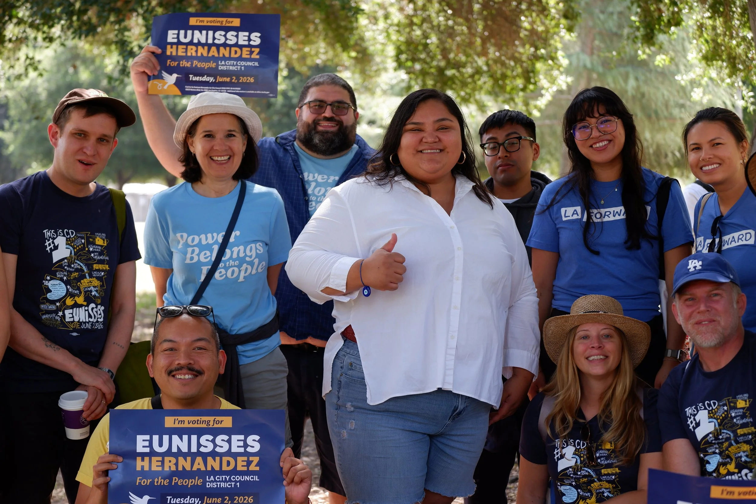 Group of diverse people at a rally or campaign event, some holding signs supporting Eunisses Hernandez for LA City Council District 1, with trees and sunlight in the background.