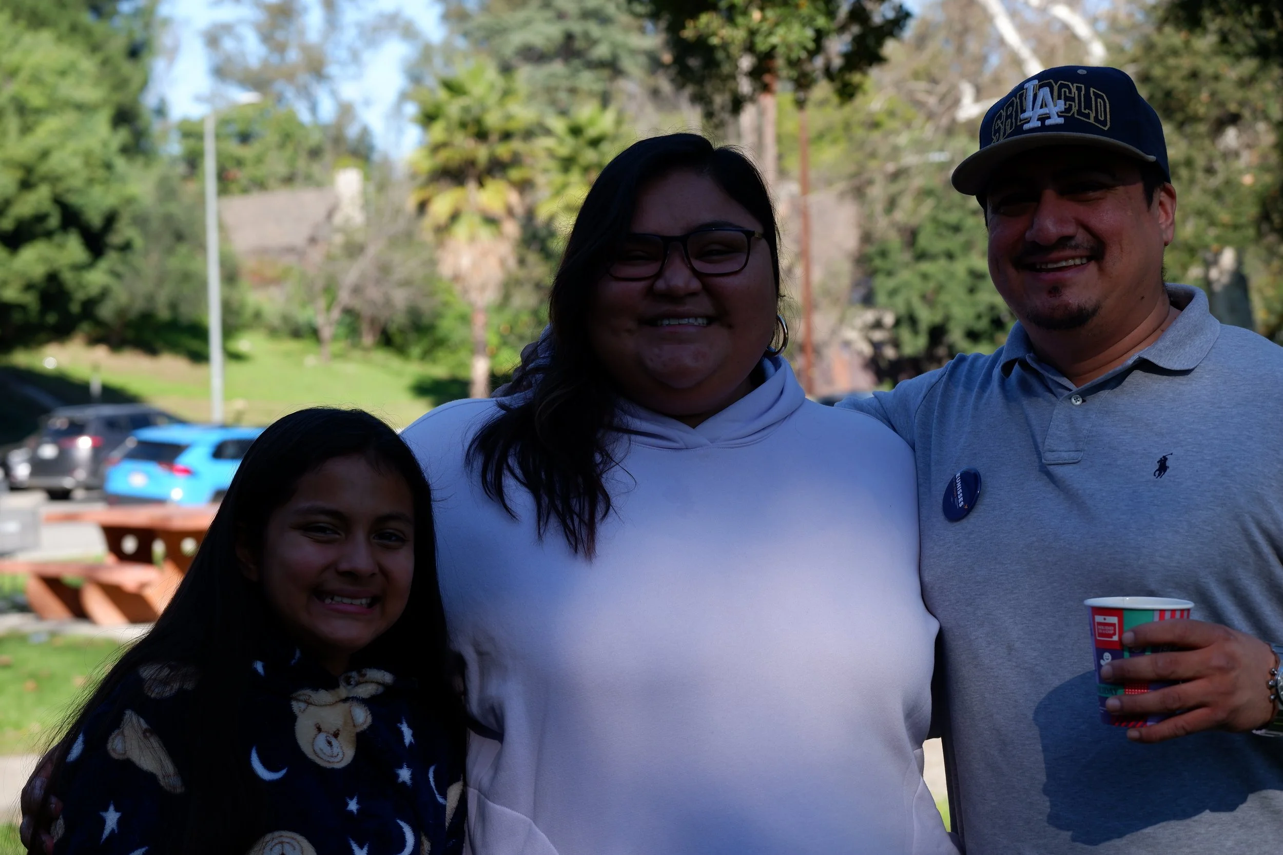 Three people, two women and one man, smiling outdoors with green trees and parked cars in the background.