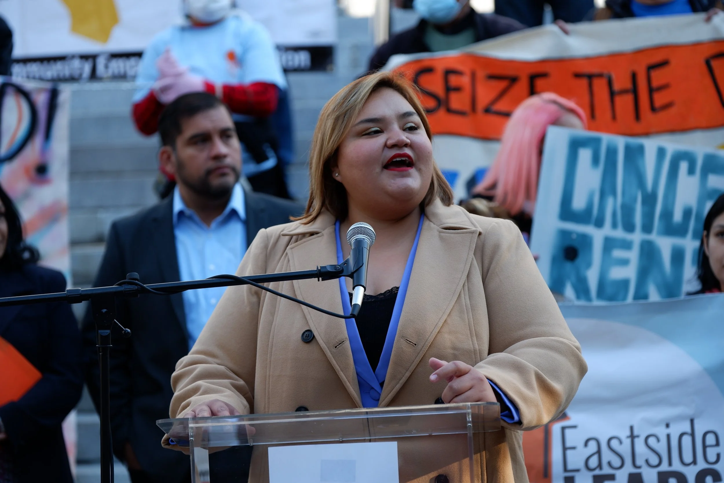 A woman speaking at a podium during a protest or rally, with a microphone in front of her and people holding signs in the background.