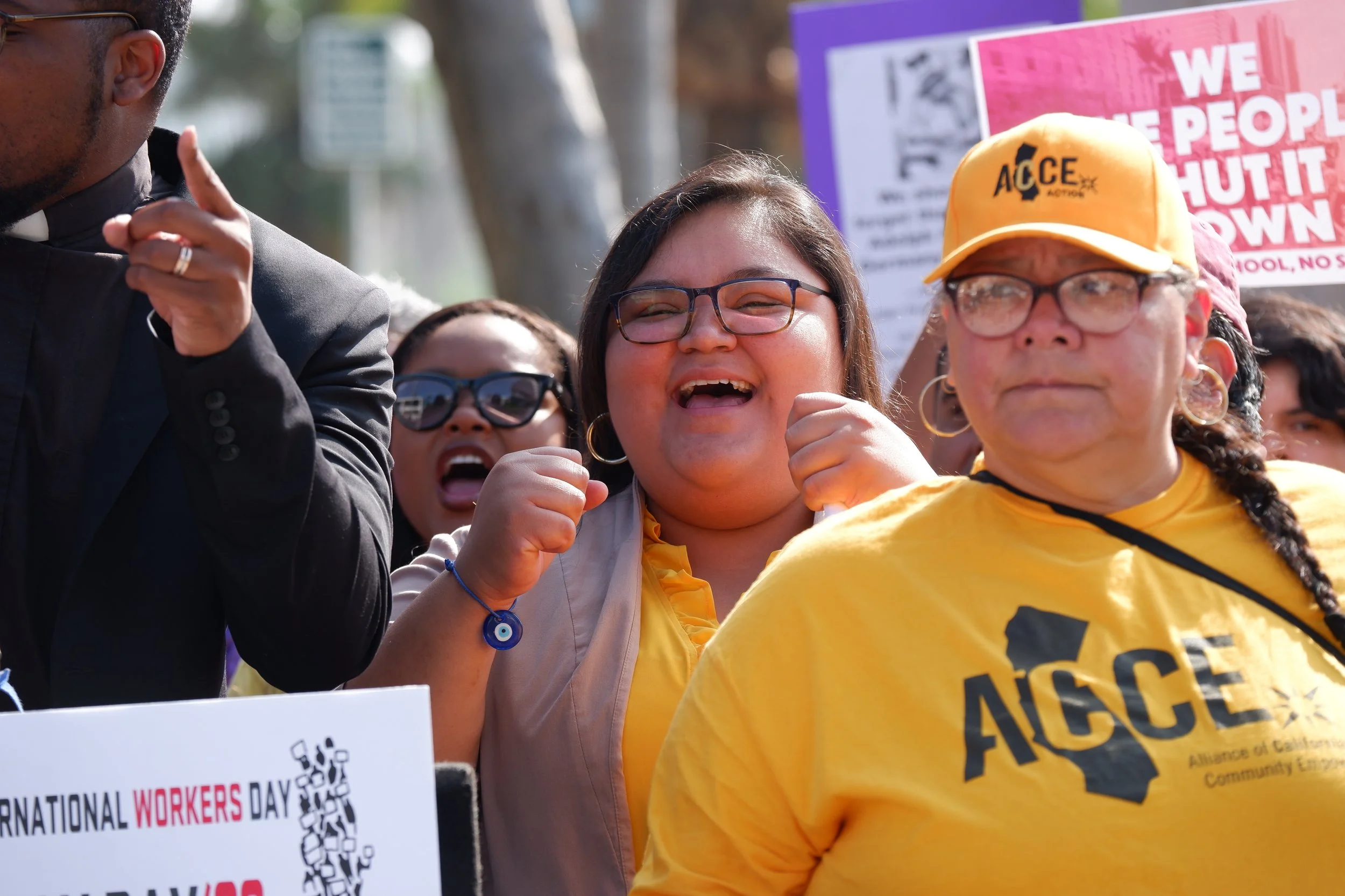 People participating in a protest or rally, holding signs about workers' rights and social justice, including a sign that reads 'NATIONAL WORKERS DAY' and one with the phrase 'WE THE PEOPLE SHUT IT DOWN' with diverse individuals smiling and engaging