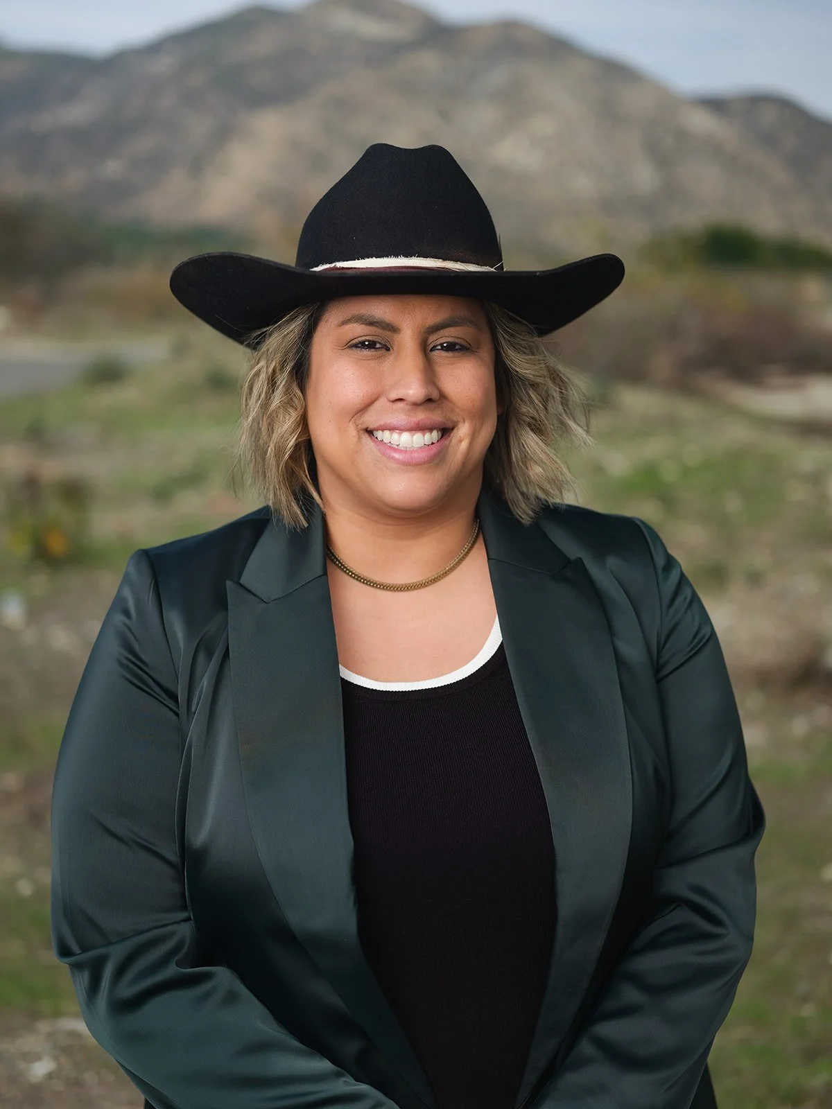 Endorsement of Caroline Menjivar for Eunisses Hernandez. She is wearing a dark green blazer, black top, and a wide-brimmed black hat in front of a green mountainous landscape.