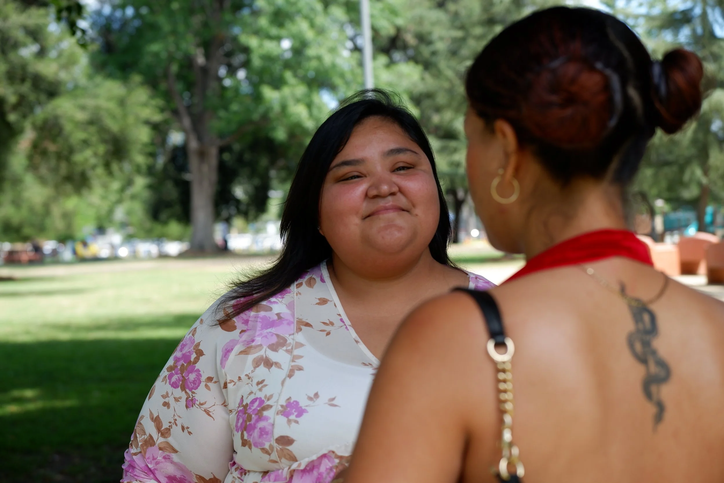 Two women conversing in a park with lush green trees in the background. One woman has long black hair, a floral blouse, and a warm smile, while the other has her back to the camera, with red-dyed hair in buns and tattoos visible.