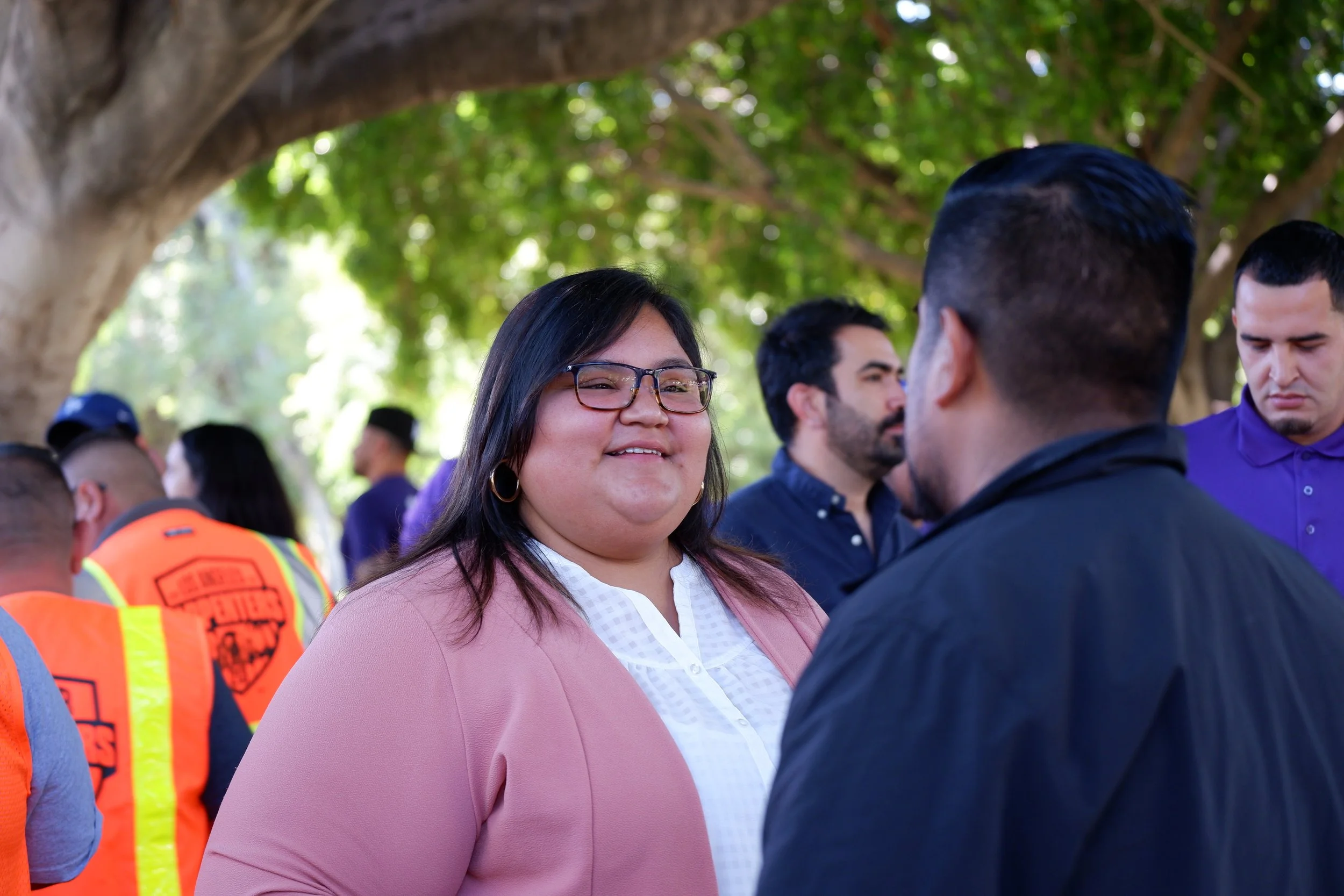 A group of diverse people gathered outdoors under a large tree, engaging in conversation. The focus is on a woman with glasses, dark hair, and a pink blazer, smiling and talking to a man with dark hair and a dark jacket. Other individuals are visible in the background, some wearing purple shirts and bright orange safety vests, indicating a community or organized event.