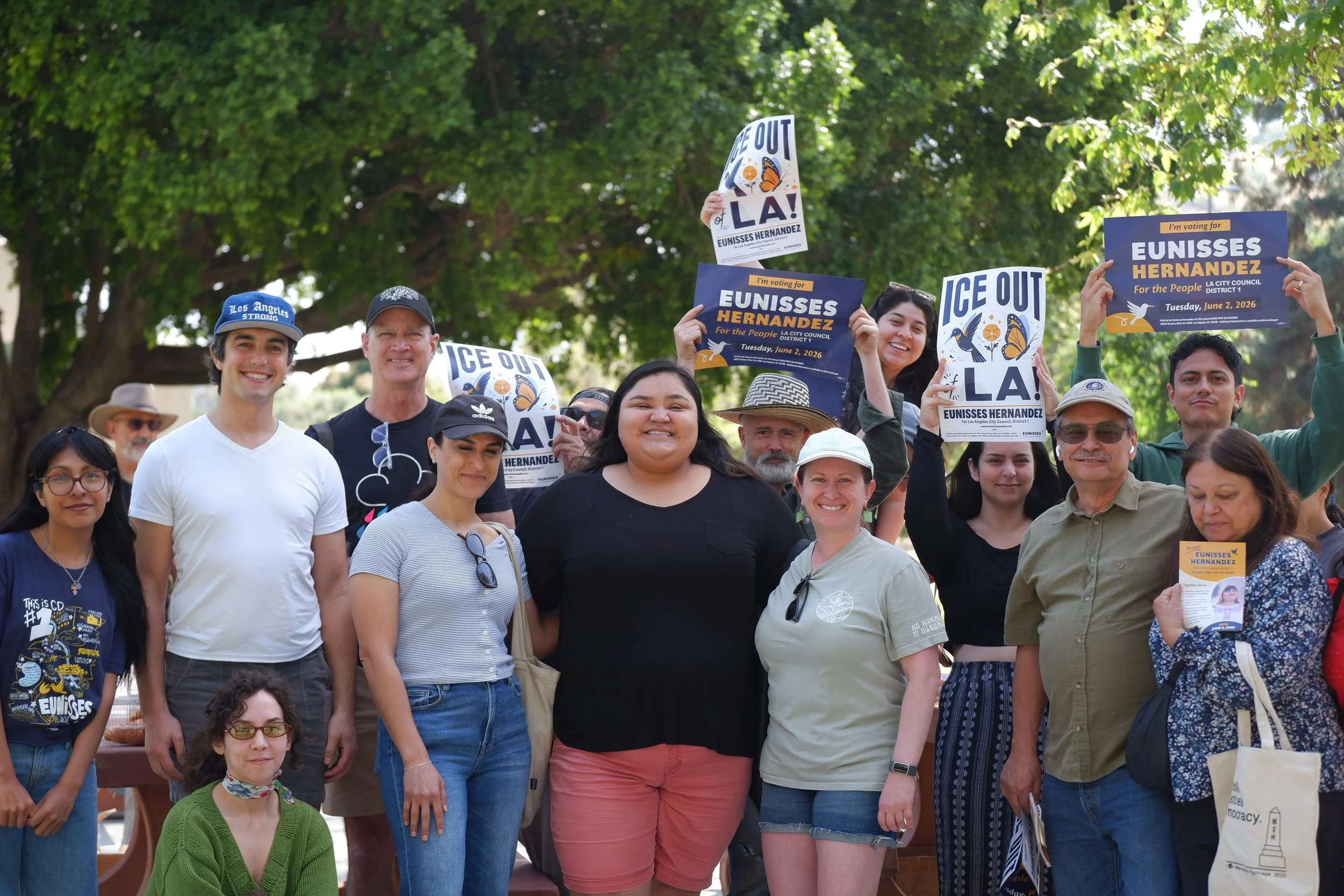 A diverse group of people gathered outdoors under trees, holding signs supporting Eunisses Hernandez for LA City Council District 1, with messages like 'ICE OUT LA!' and 'I'm voting for Eunisses Hernandez,' during a community event or rally.