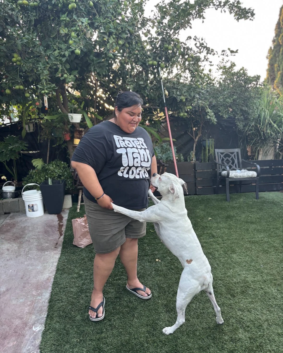 A woman wearing a black T-shirt with white text and khaki shorts is playing with a large white dog in a backyard. The dog is standing on its hind legs with paws on the woman's waist. The backyard has green grass, potted plants, and trees with apples. In the background, there are black garden planters and a bench.