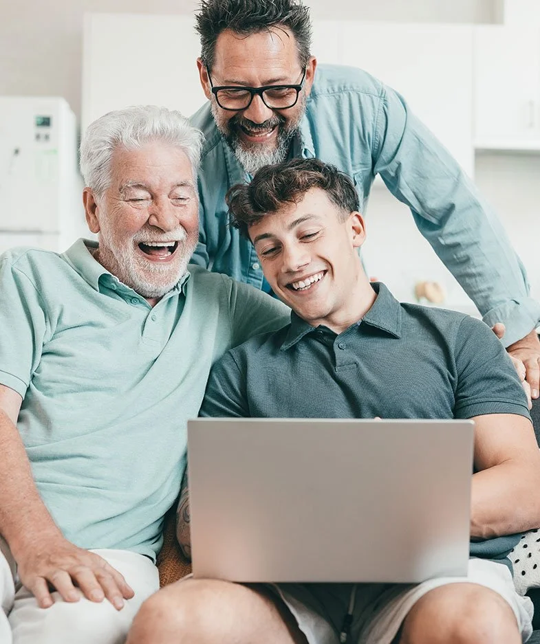 Three generations of men smiling and looking at a laptop together in a cozy home environment.
