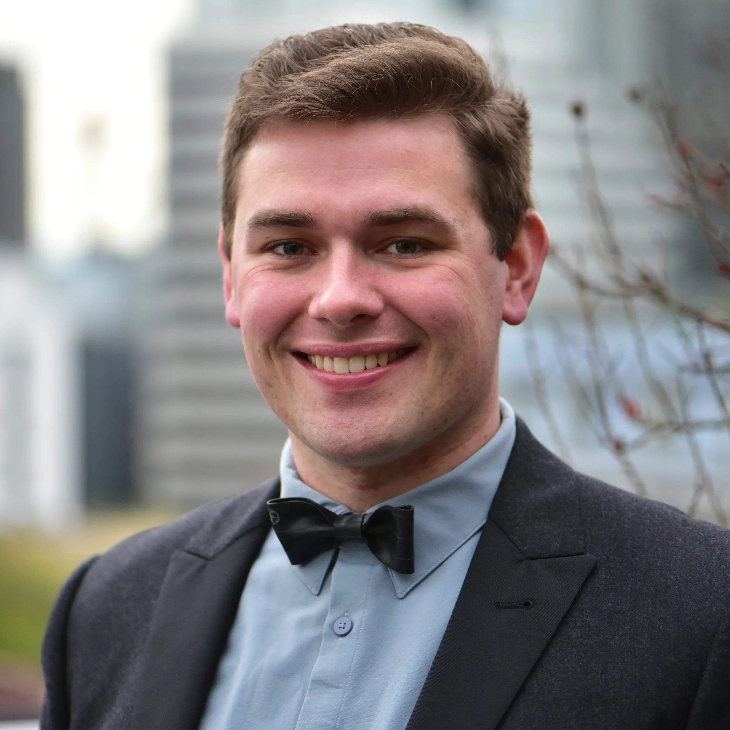 Dr. Philip Walczak, wearing a formal suit, light blue shirt, and black bow tie, smiling outdoors with city buildings and blurred tree branches in the background.