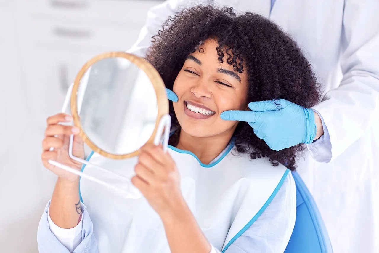 A woman smiling while examining her teeth in a mirror at a dental appointment, with a dentist gently touching her cheek.