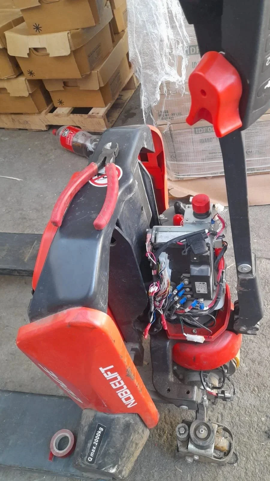 Montacargas con componentes internos visibles, en un almacén con cajas de cartón y una botella de Coca-Cola en el fondo.