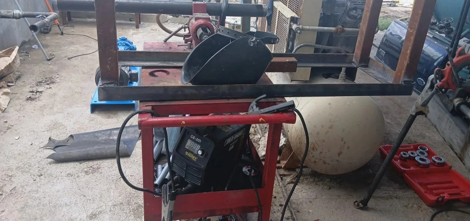 Área de taller con equipo y herramientas de metalurgia, incluyendo una mesa, una máquina soldadora, partes metálicas, y accesorios diversos en un espacio de trabajo industrial.