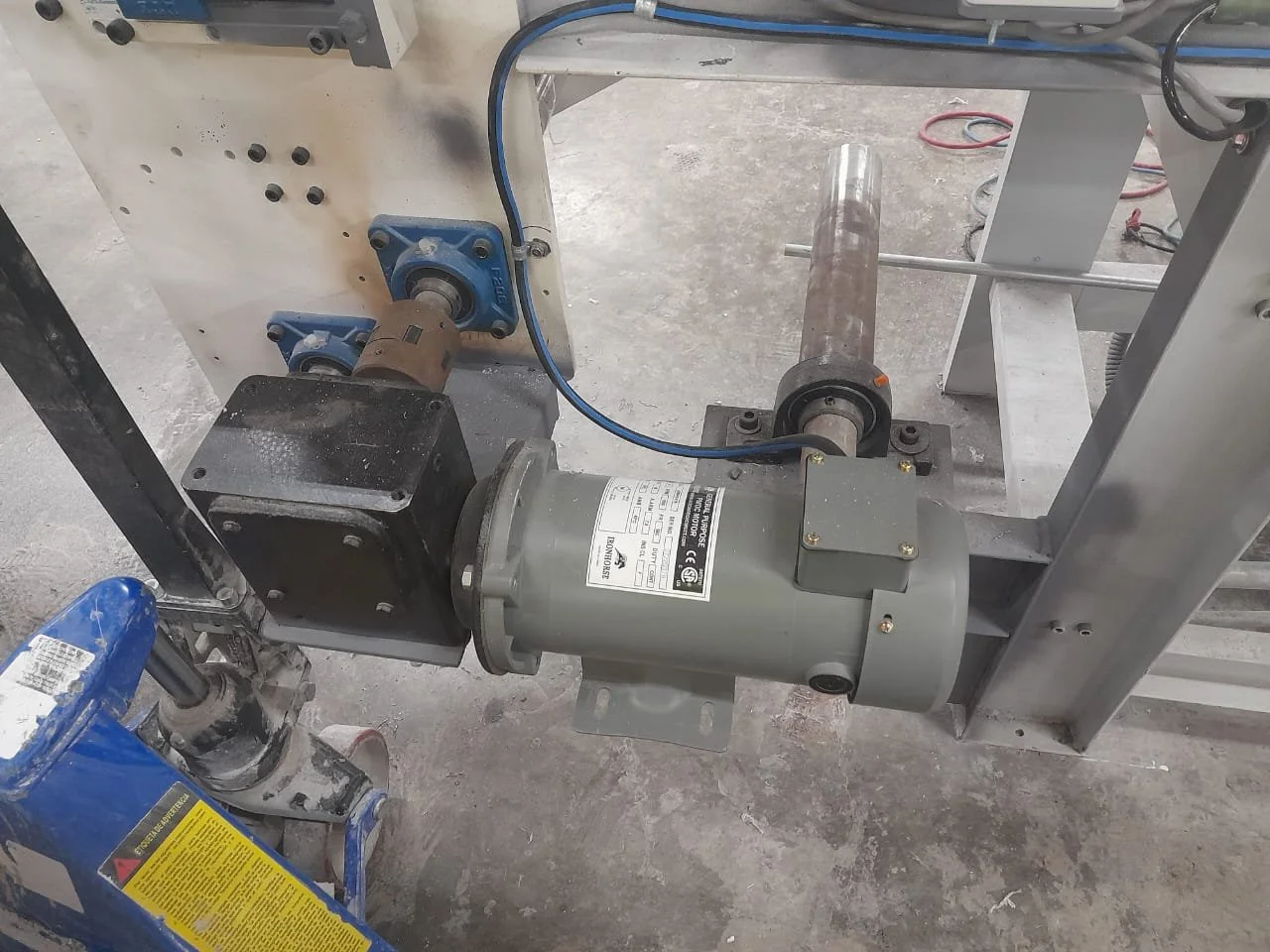 Componentes mecánicos y eléctricos en una máquina industrial, incluyendo un motor, rodamientos, y un cilindro de metal en un ambiente de taller.