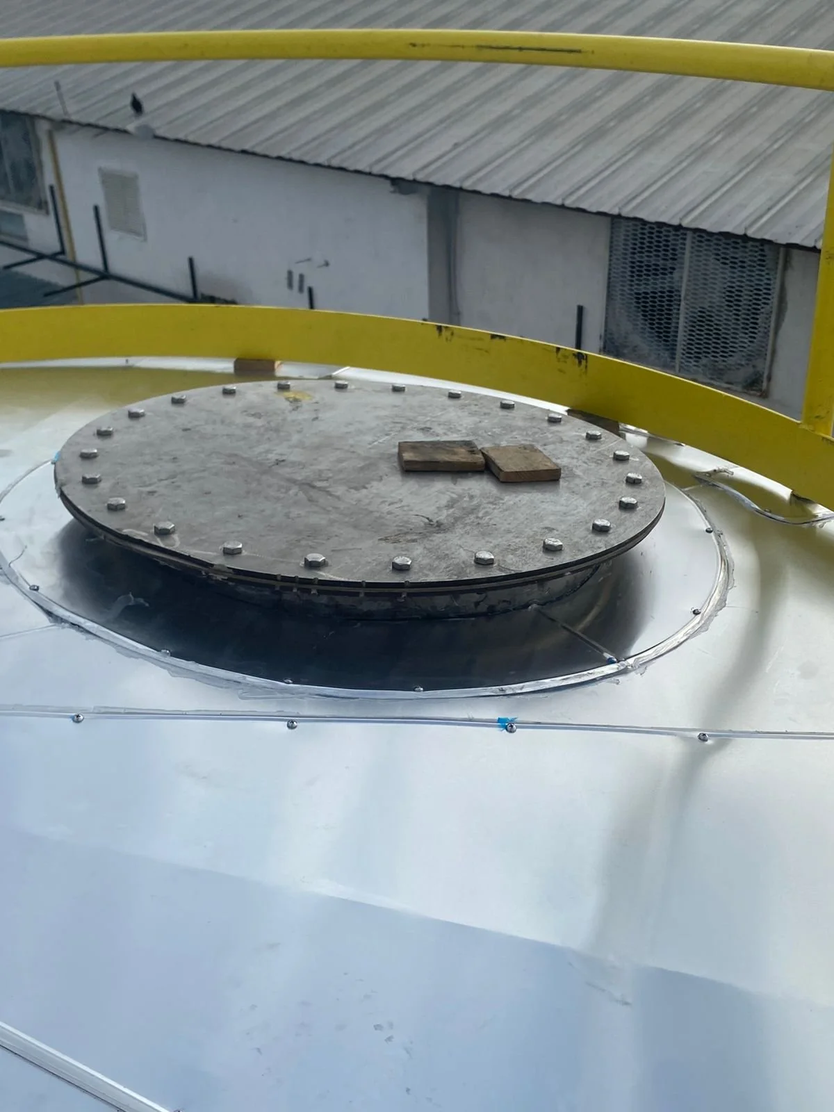 Tapa metálica en la parte superior de una estructura industrial, rodeada de barras amarillas, en un entorno urbano.