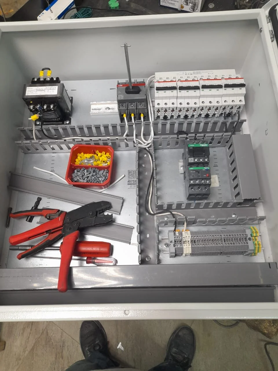 Caja de distribución eléctrica con componentes y herramientas, incluyendo un cable corto, pinzas, tornillos, y bloques de conexión.