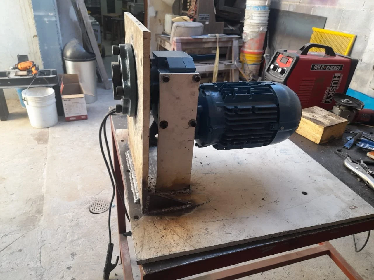 Motor eléctrico montado en una estructura de madera en un taller de carpintería, con herramientas y materiales de construcción en el fondo.