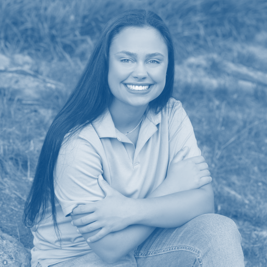 A woman smiling, sitting outdoors with arms crossed, in front of a grassy background.