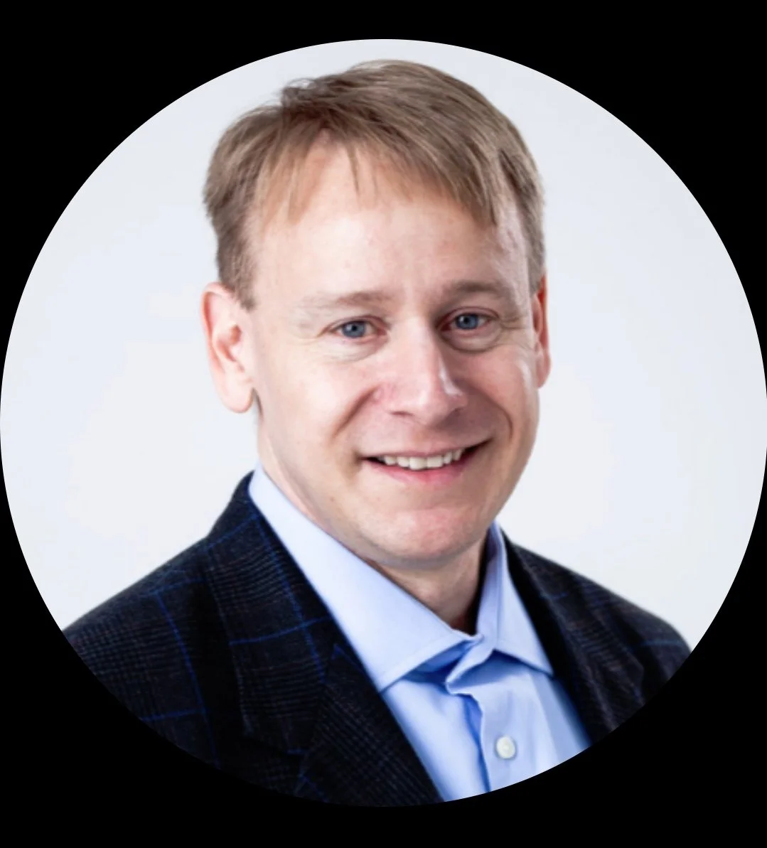 Headshot of a smiling man with red hair, wearing a blue shirt and a dark blazer with a subtle plaid pattern, against a white background.