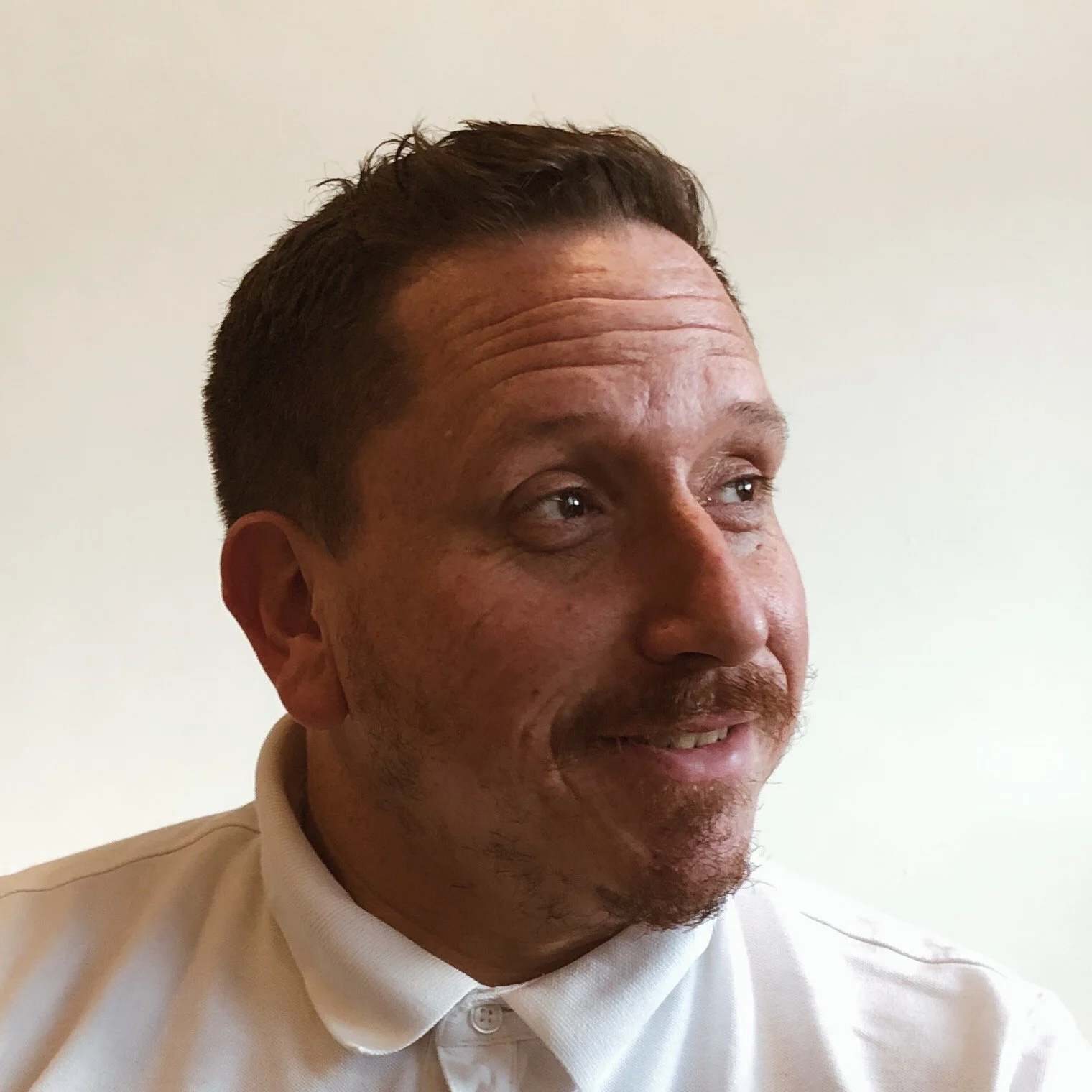Close-up of a middle-aged man with short dark hair, wearing a white collared shirt, looking to his right with a slight smile, against a plain light background.