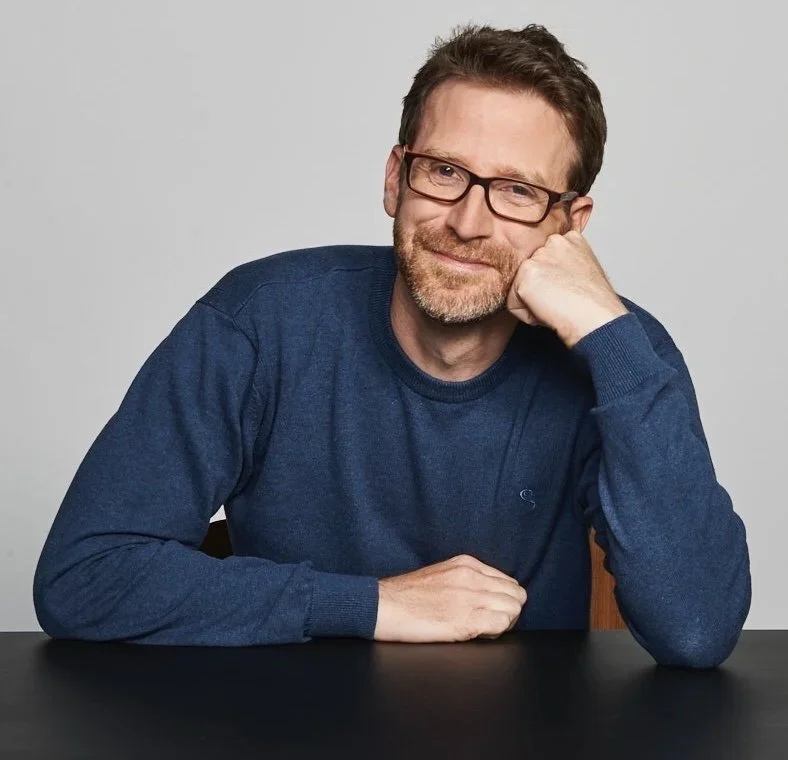 A man with glasses and a beard wearing a blue sweater, resting his head on his hand, sitting at a black table against a plain gray background.