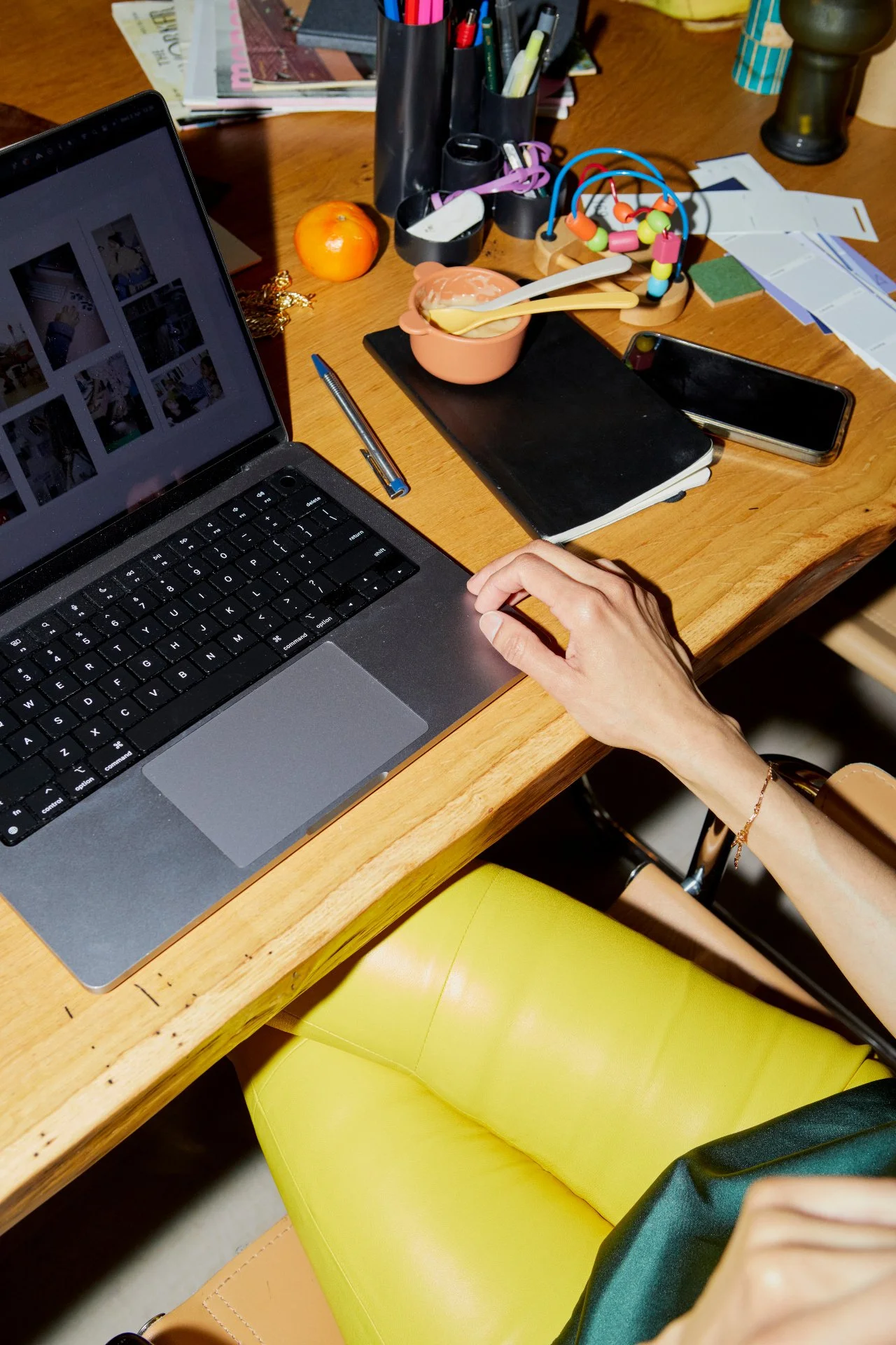 A cluttered desk with a laptop, smartphone, notebooks, a pen, a bowl with spoons, a toy bead maze, an orange, and various papers and office supplies. A person is using the laptop, with their hand on the trackpad, and is sitting on a yellow chair.