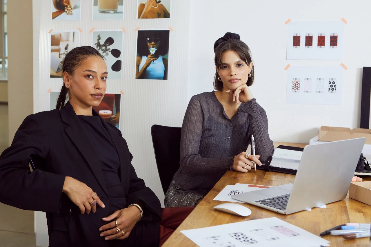 Two women sitting at a wooden table in an office, with a laptop, notebooks, and pens. One woman has a hand on her hip, and the other is resting her chin on her hand, looking at the camera. The background features photos and design sketches taped to the wall.