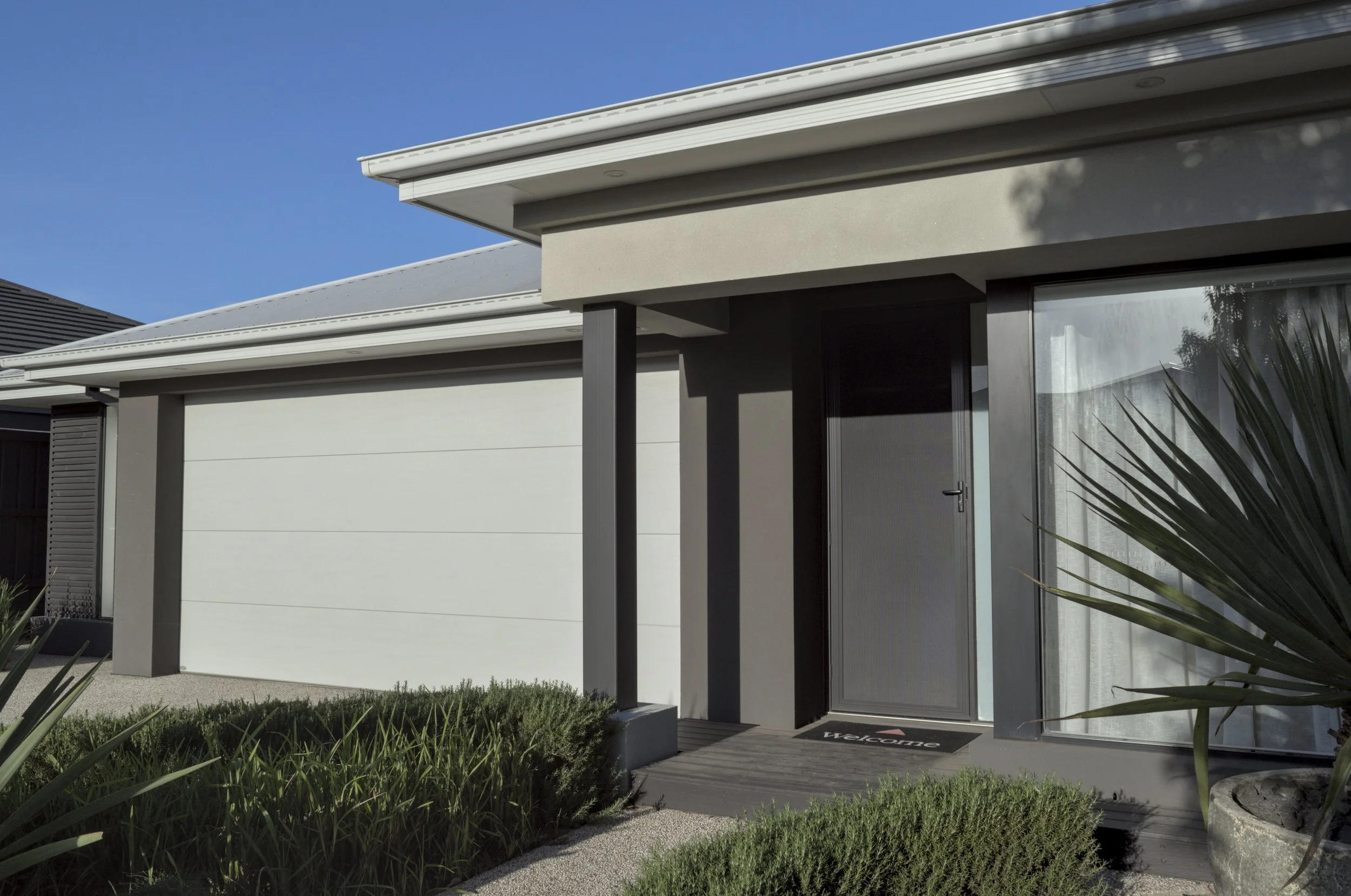 A dark gray textured vase with green and reddish plant stems, a white cylindrical device with a metallic base, and two books titled "The Wellness Cabinet" and "Plants at Your Place" arranged on a white surface against a plain light wall.
