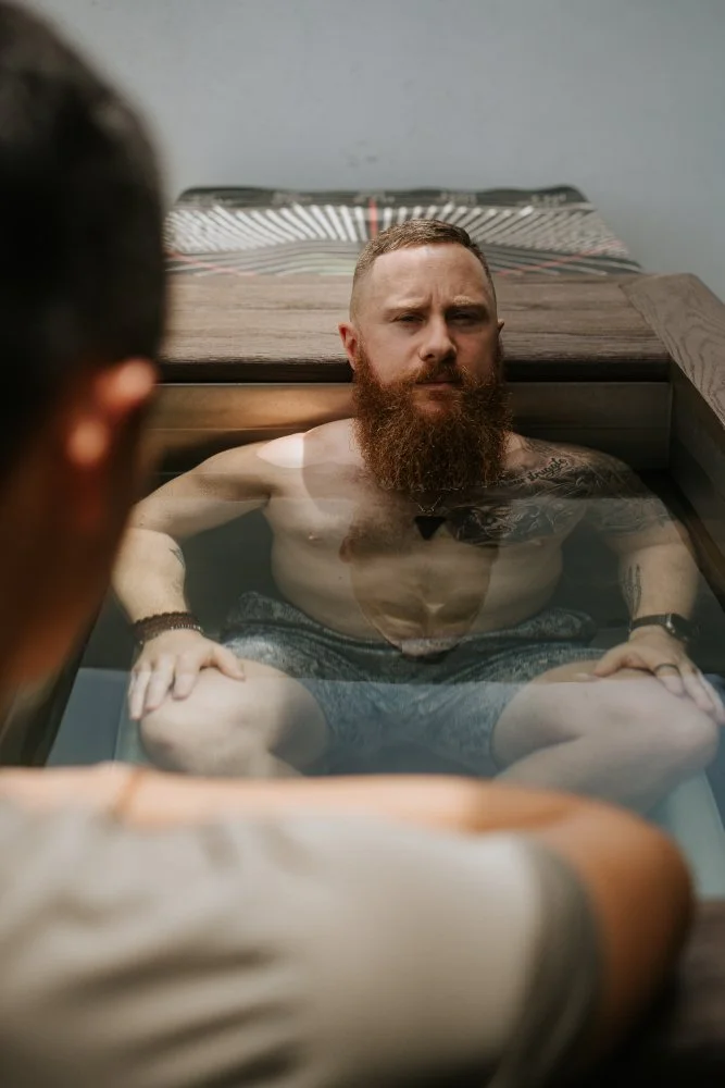 A man with a beard and tattoos sitting in a foot bath or immersion tub, looking at the camera with a serious expression.