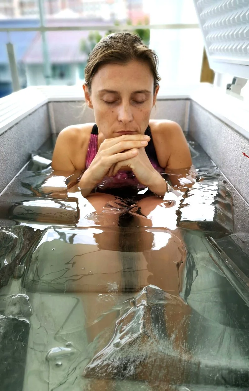 A woman lying in a cold plunge, viewed from above, with feet immersed in water, inside a wooden frame structure.