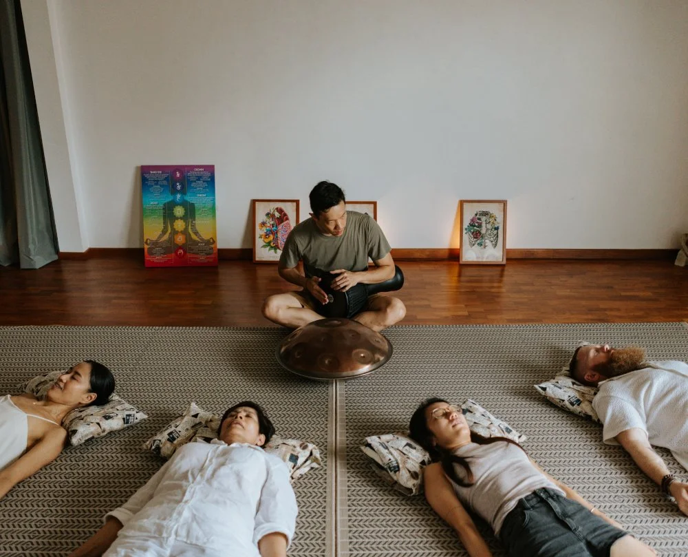 A sound healing session with five people lying on mats on the floor, a man seated cross-legged in the center playing a handpan, and colorful artwork and posters against the wall in a spacious room.