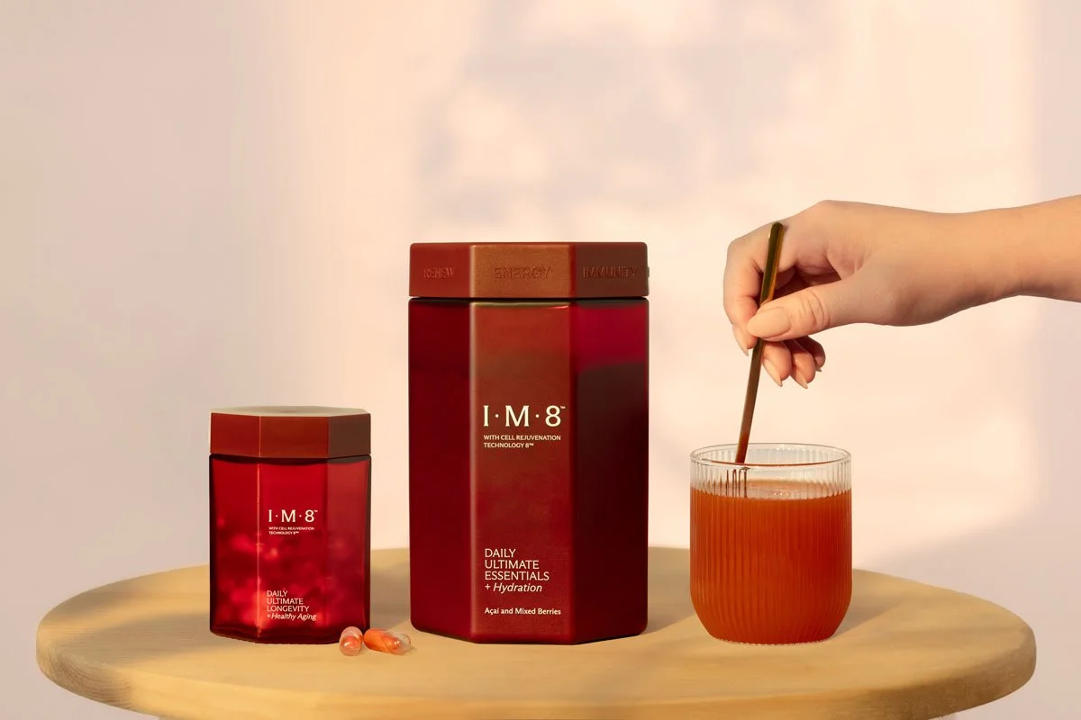 Two red boxes labeled I·M·8 with different sizes and a glass of orange-red drink with a hand stirring it using a golden spoon on a wooden surface.