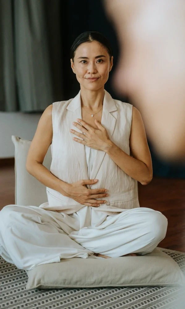 A woman practicing yoga indoors, sitting cross-legged on a cushion with her hand on her chest and stomach, in a meditative posture.