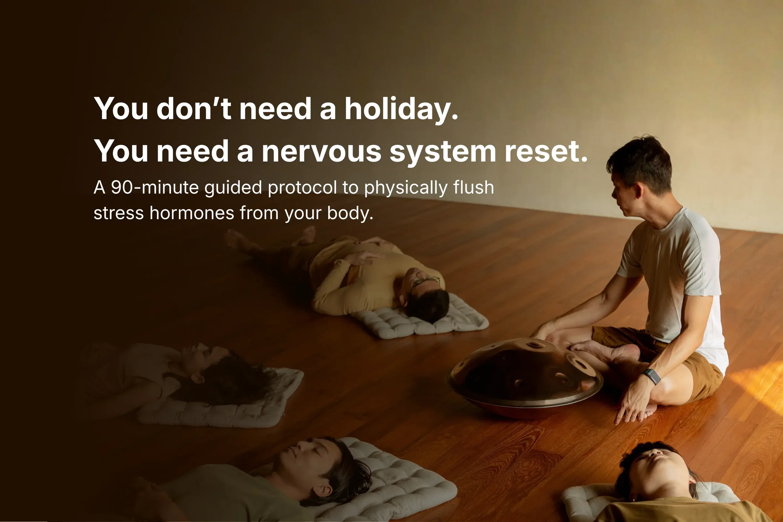 A group of people lying on mats in a tranquil studio during a 90-minute nervous system reset protocol, with a facilitator playing a handpan drum in the foreground.