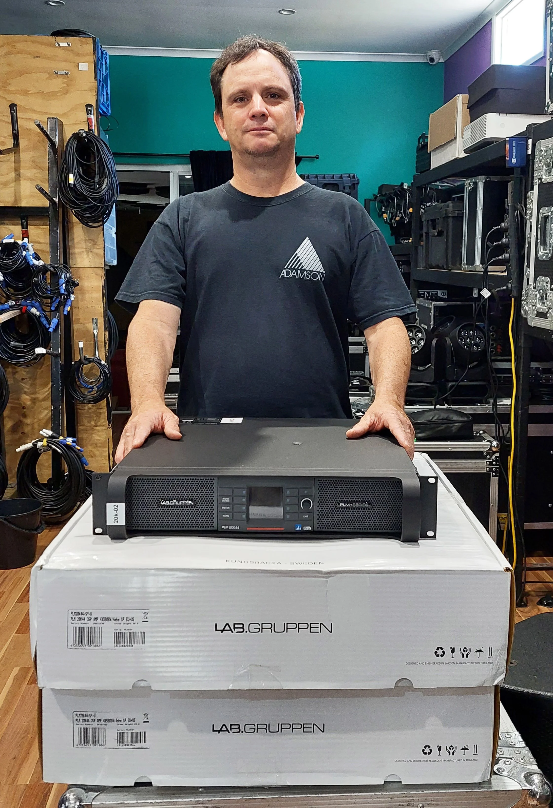 Man standing behind a large audio amplifier on a white LAB.GRUppen box, surrounded by audio equipment and cables in a workshop.