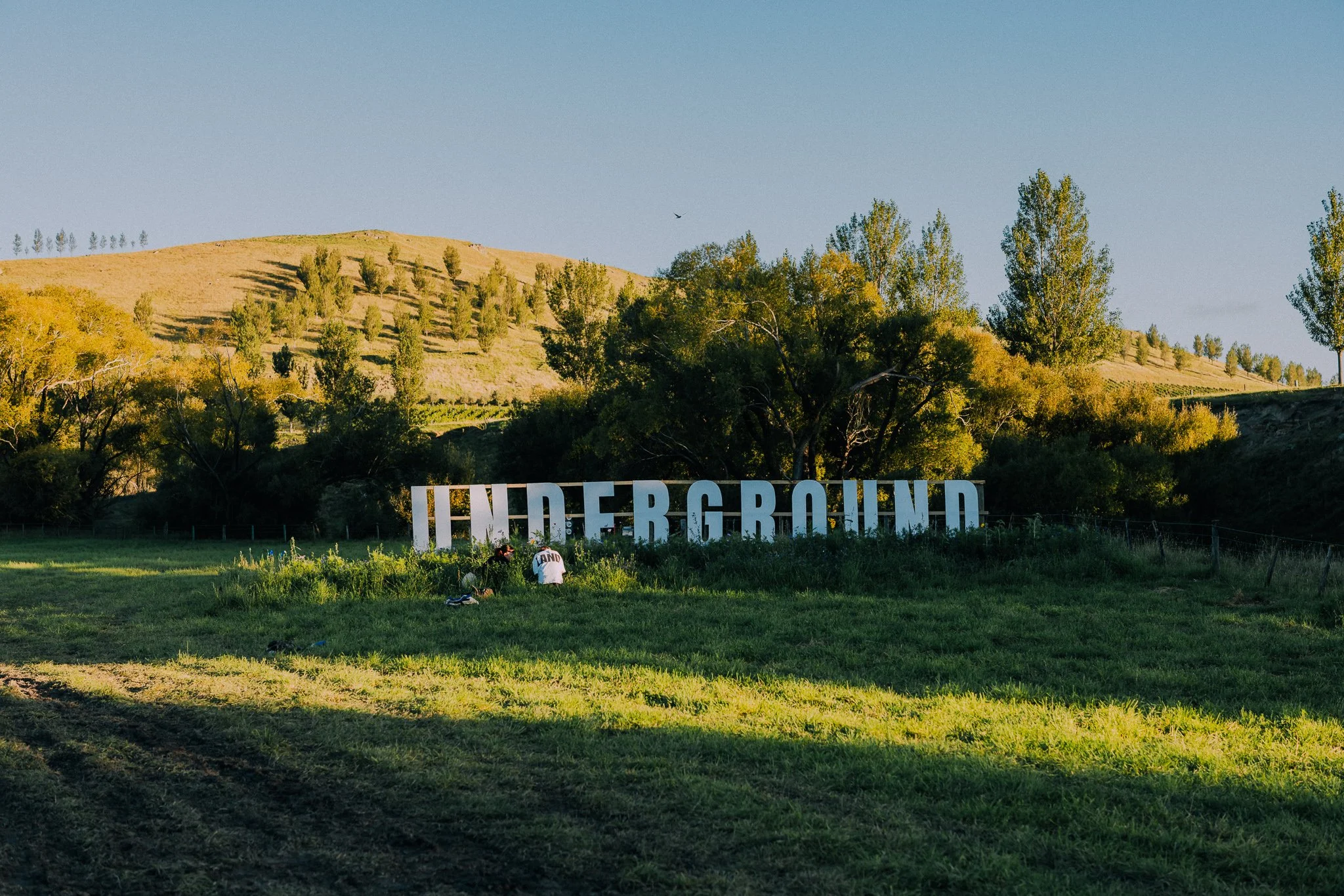 Large white letters spelling 'UNDERGROUND' on a grassy field with trees and hills in the background.