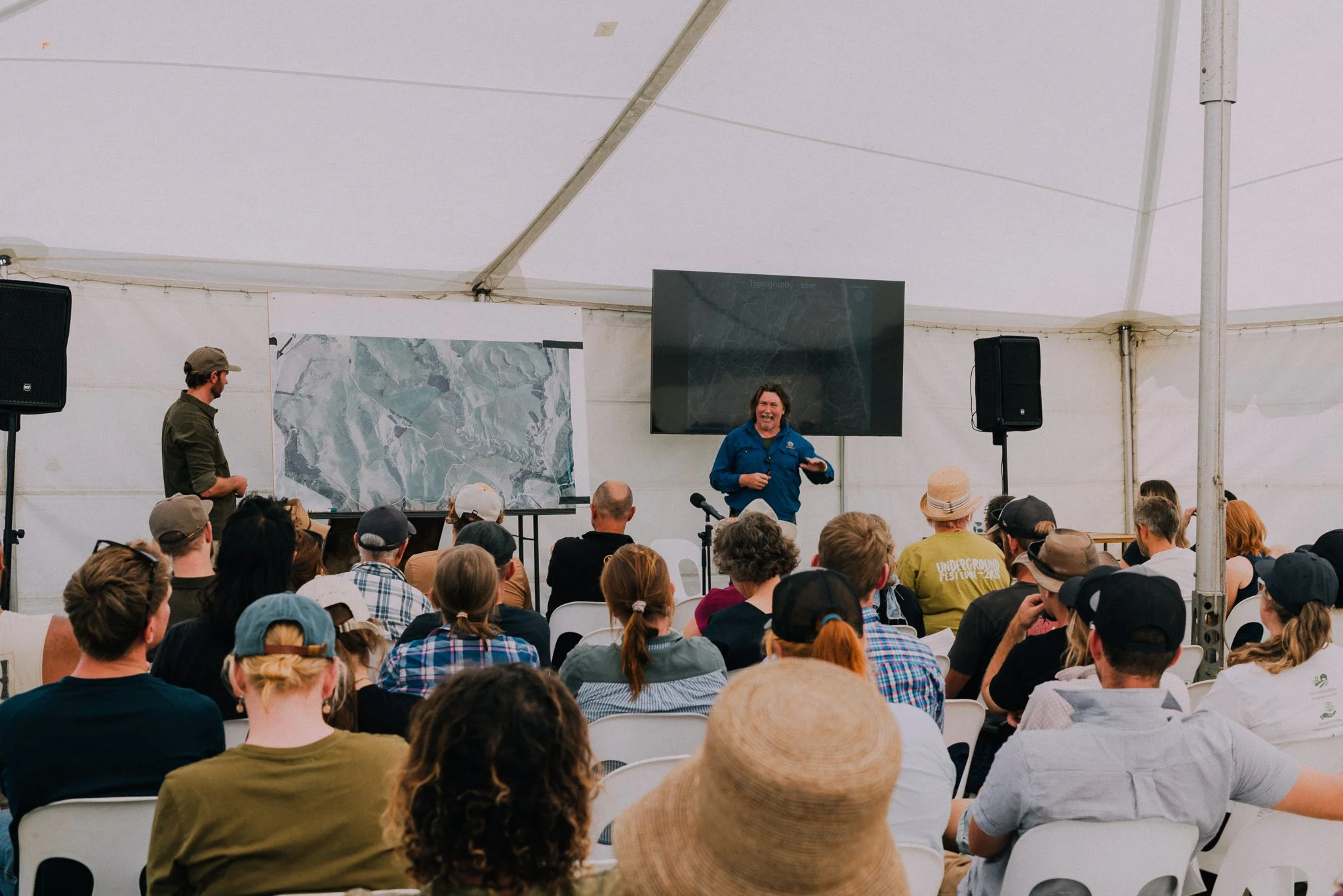 A presenter stands in front of an audience inside a large white tent, giving a presentation. There is a large screen and a map on a board behind the presenter. The audience is seated on white chairs, and some people are wearing hats.