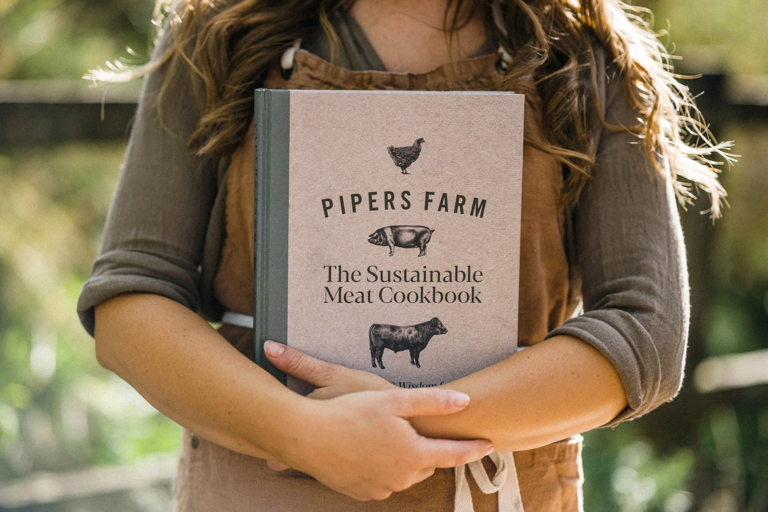 Person holding a cookbook titled 'Pipers Farm: The Sustainable Meat Cookbook' with illustrations of a chicken, pig, and cow on the cover.