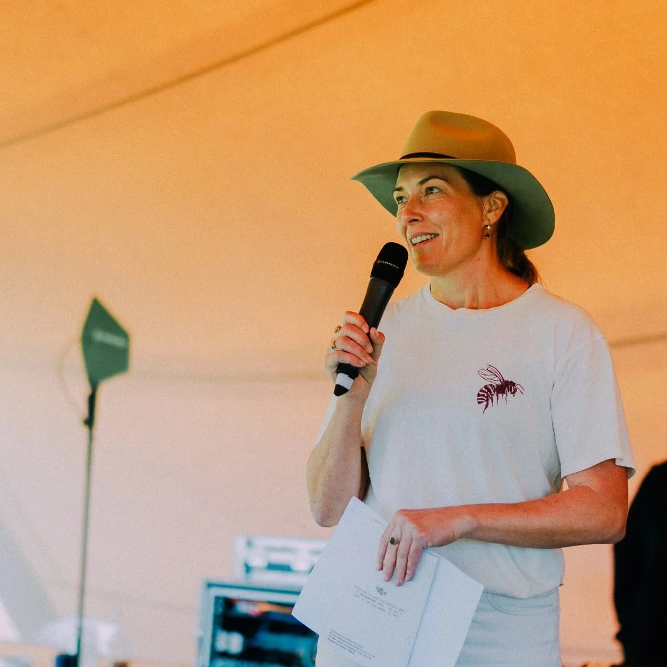 A woman wearing a gray wide-brim hat and a white t-shirt with a bee logo, holding a microphone and a sheet of paper, speaking at an event.