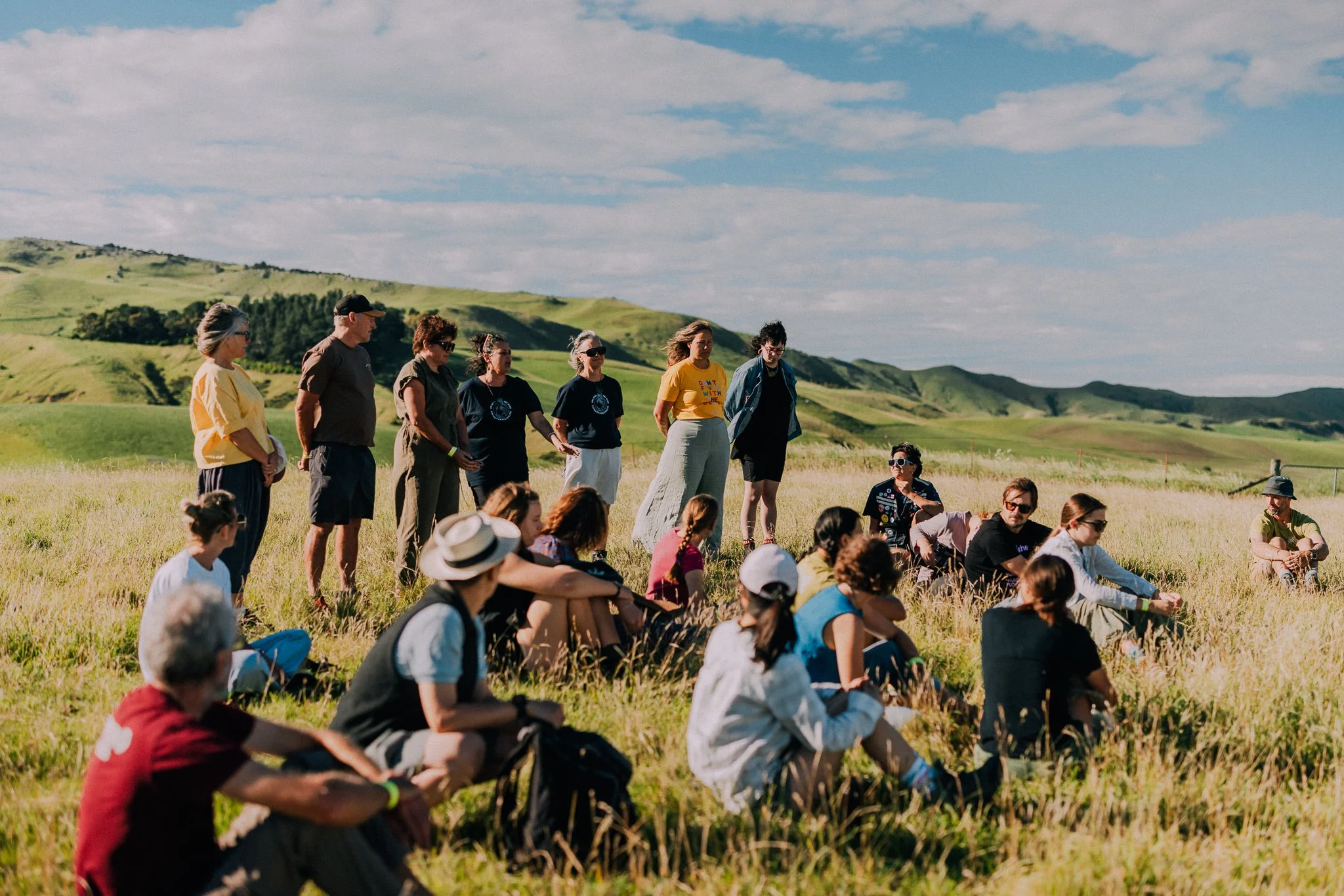 A group of people gathered outdoors in a grassy field on a sunny day, with rolling green hills and a partly cloudy sky in the background.