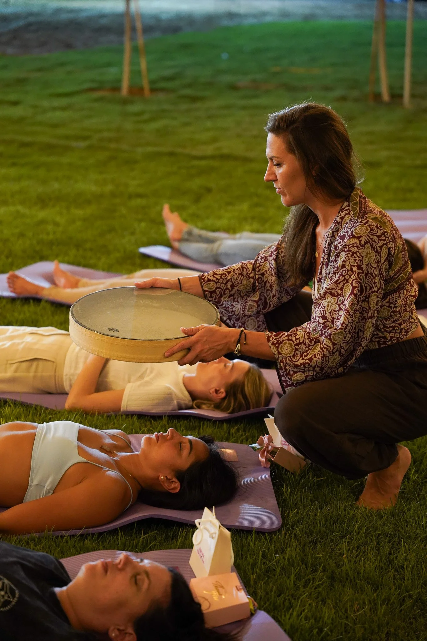 A woman is kneeling on the grass, holding a round drum over a person lying on a yoga mat. The scene is outdoors, with several people relaxing on yoga mats on the grass.