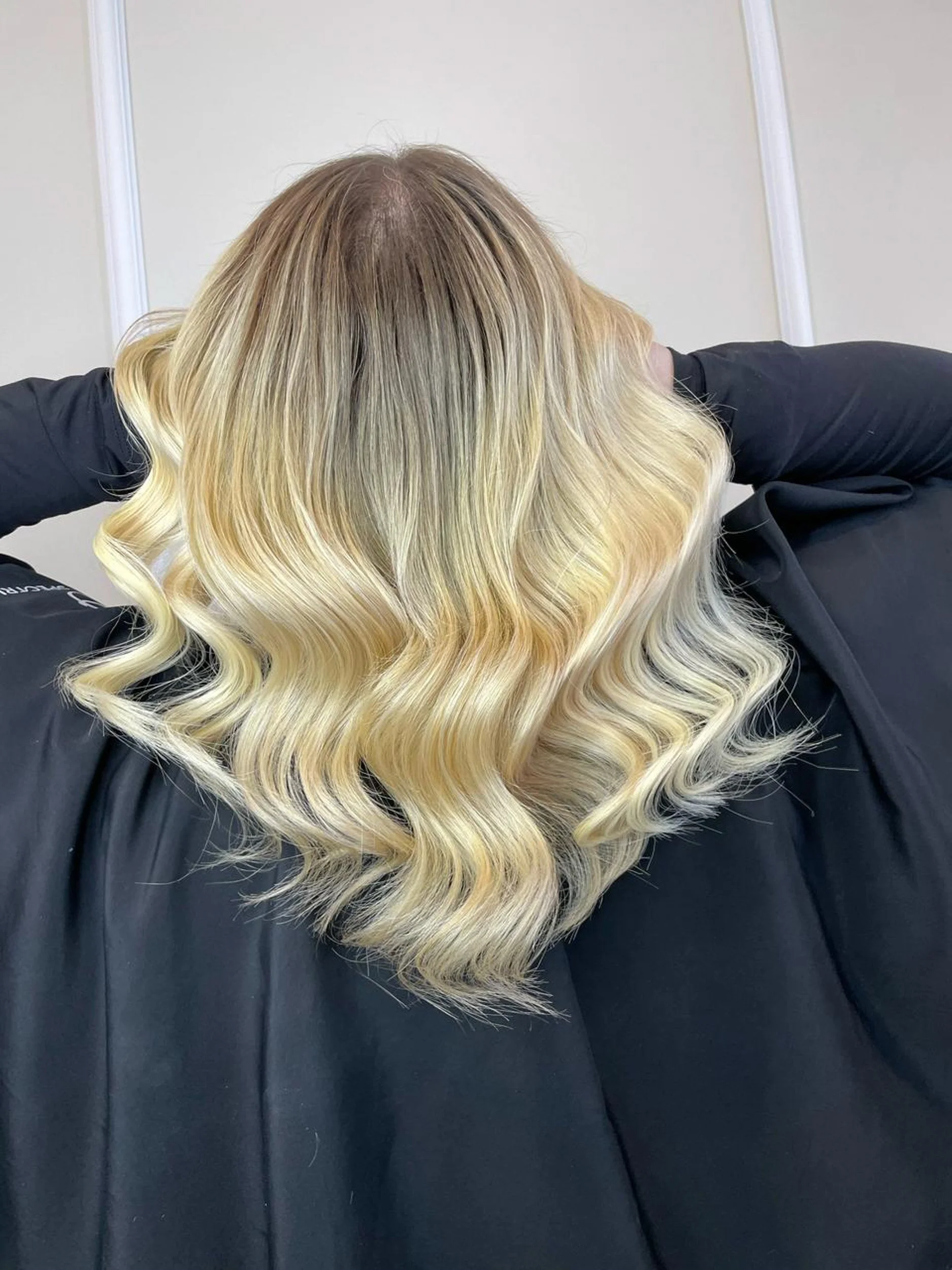 Back view of a person with wavy blonde hair, featuring dark roots, wearing a black garment.