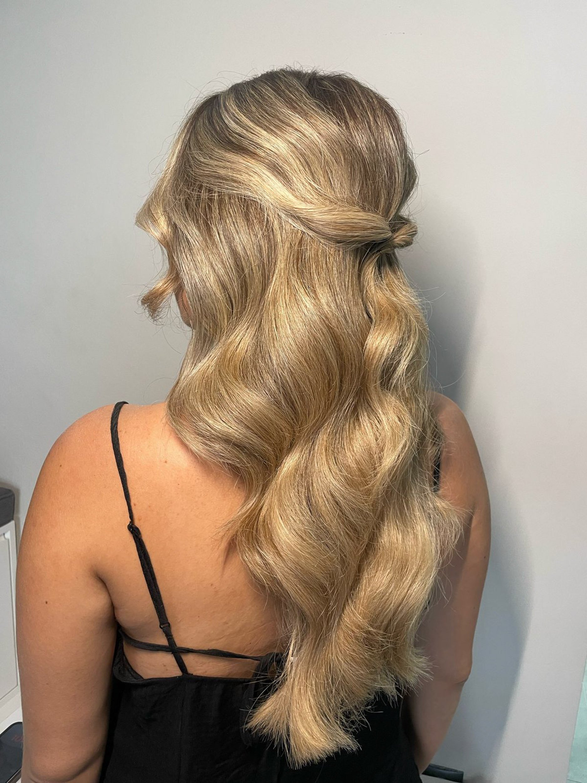 Woman with long wavy blonde hair styled in a half-up half-down knot, wearing a black spaghetti strap top.