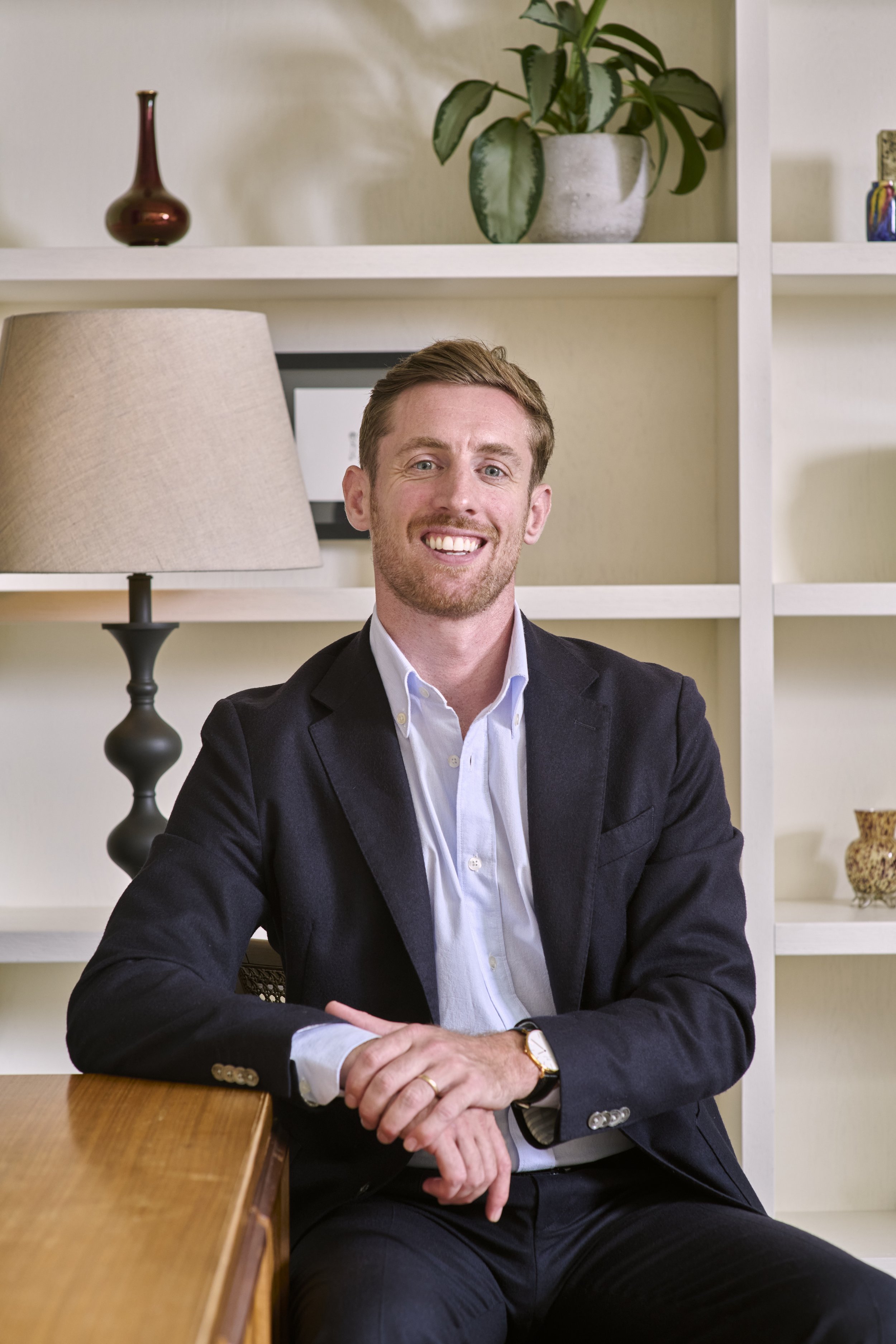 A man in a suit smiling, sitting at a wooden table with his hands clasped, in front of a white bookshelf with decorative items, a lamp, and a potted plant.