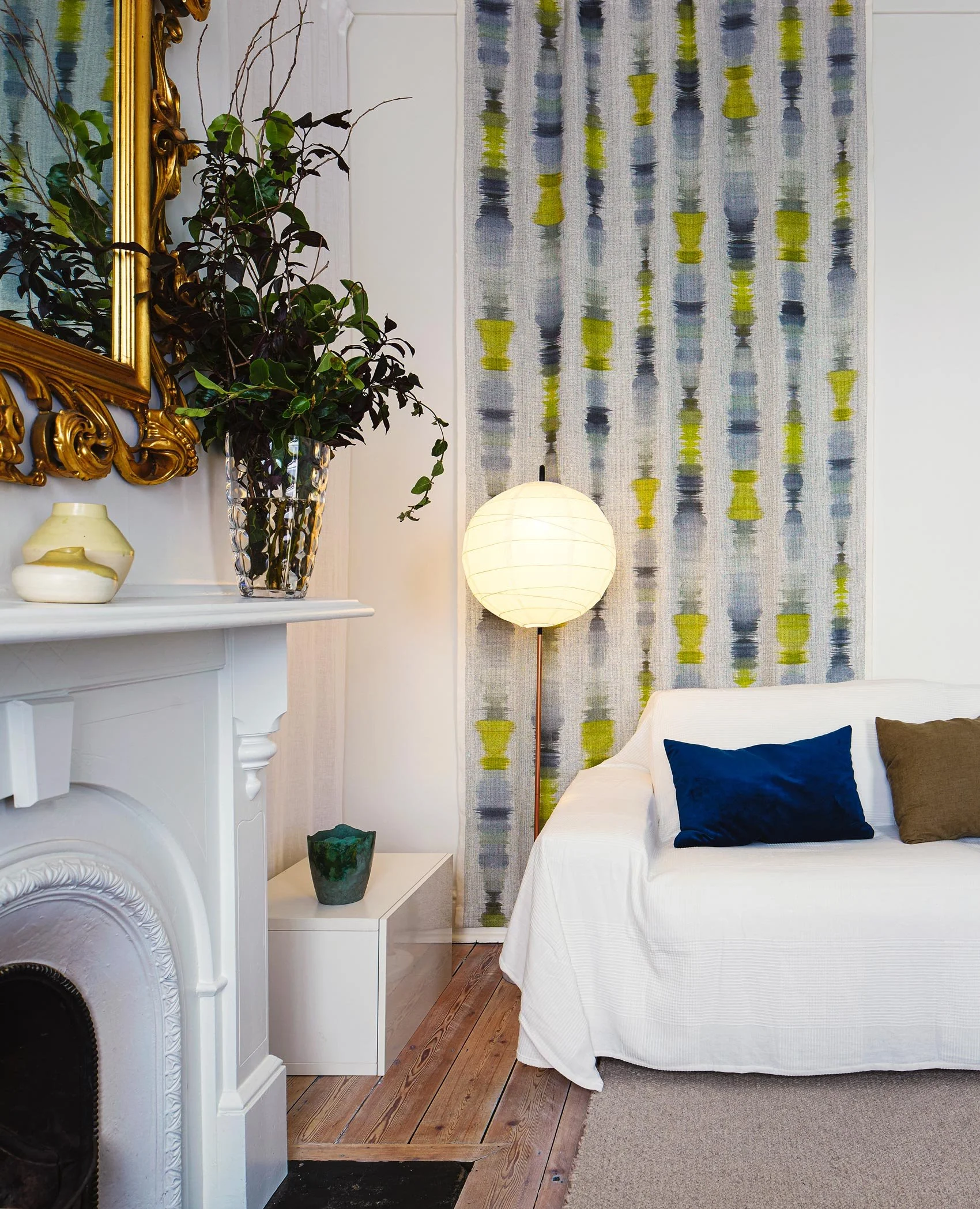 Cozy living room corner with a white sofa draped in a white cover, decorated with a navy blue and a brown pillow. A tall ovoid paper lantern stands beside the sofa, illuminating the room. Behind it, a colorful vertical patterned curtain features shades of blue, green, and gray. To the left, a white fireplace mantel holds decorative items including a small ceramic piece and a glass vase with green and dark plants, with an ornate gold-framed mirror hanging above.