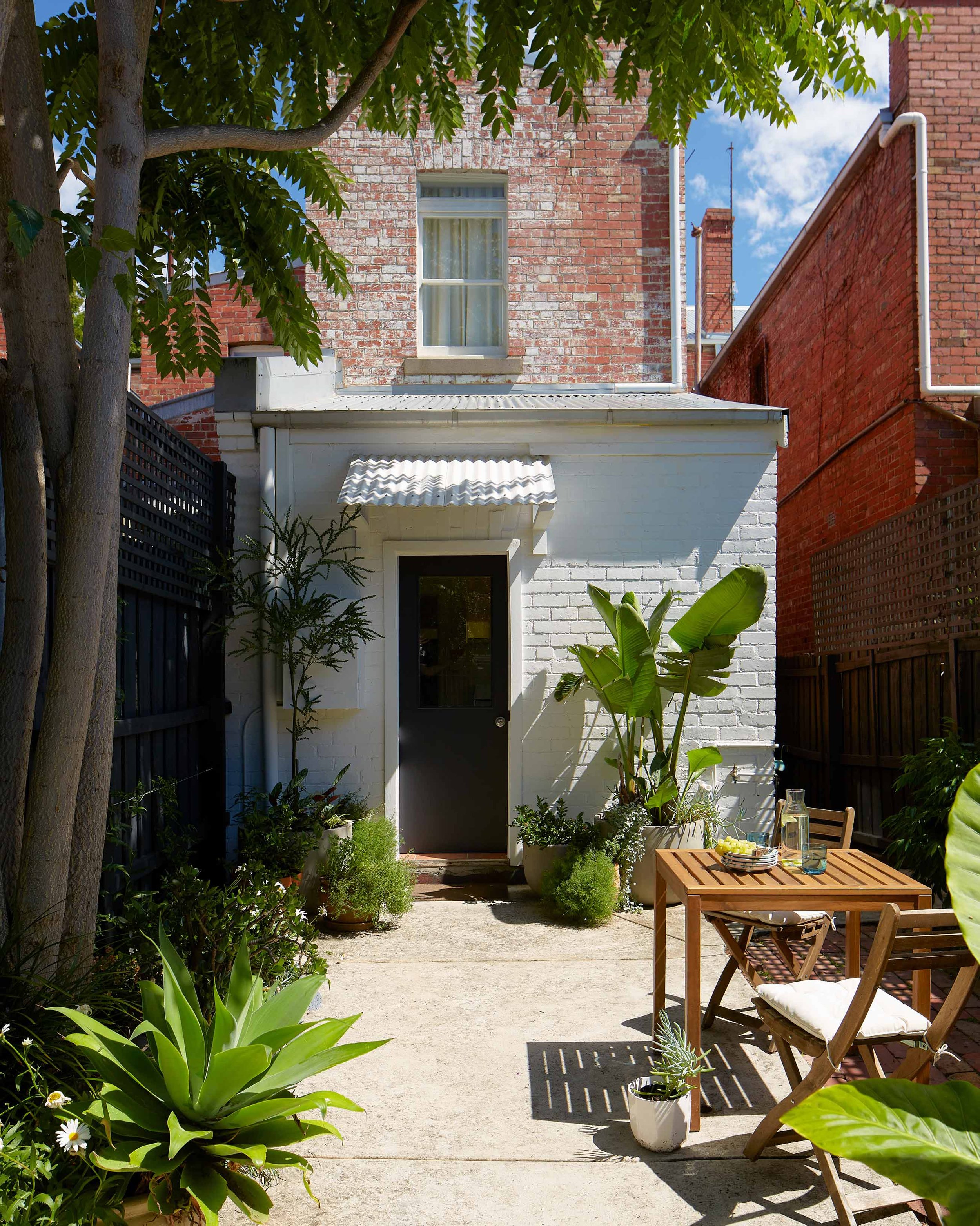 A courtyard with a black door, potted plants, a wooden table with two chairs, and lush greenery under a tree.