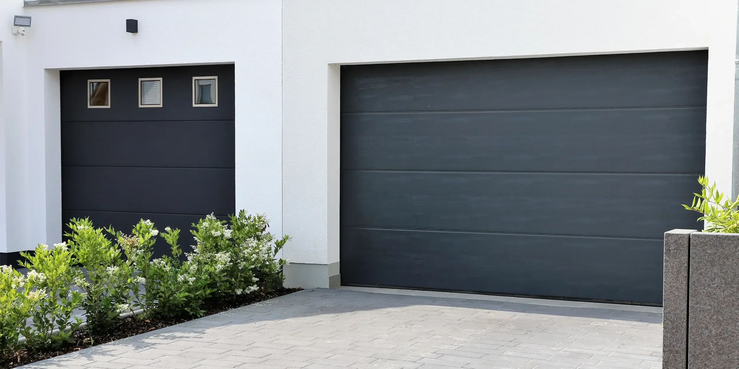 Modern house with two black garage doors, small window panes on the left garage door, white exterior walls, and landscaped greenery with white flowers and bushes in front.