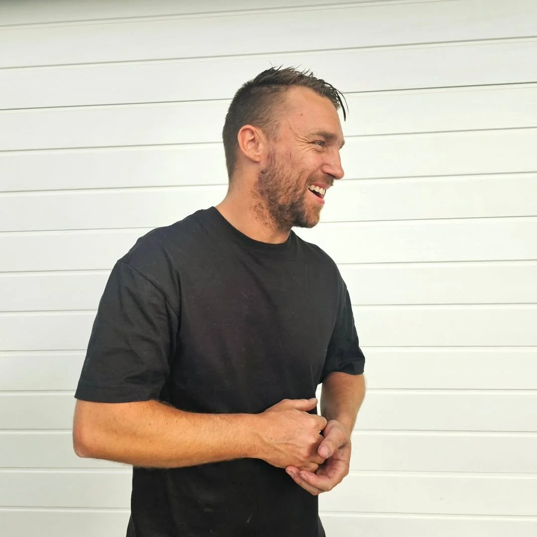 A man with short dark hair and a beard, wearing a black t-shirt, smiling and looking to the side, standing against a white wall with horizontal panels.