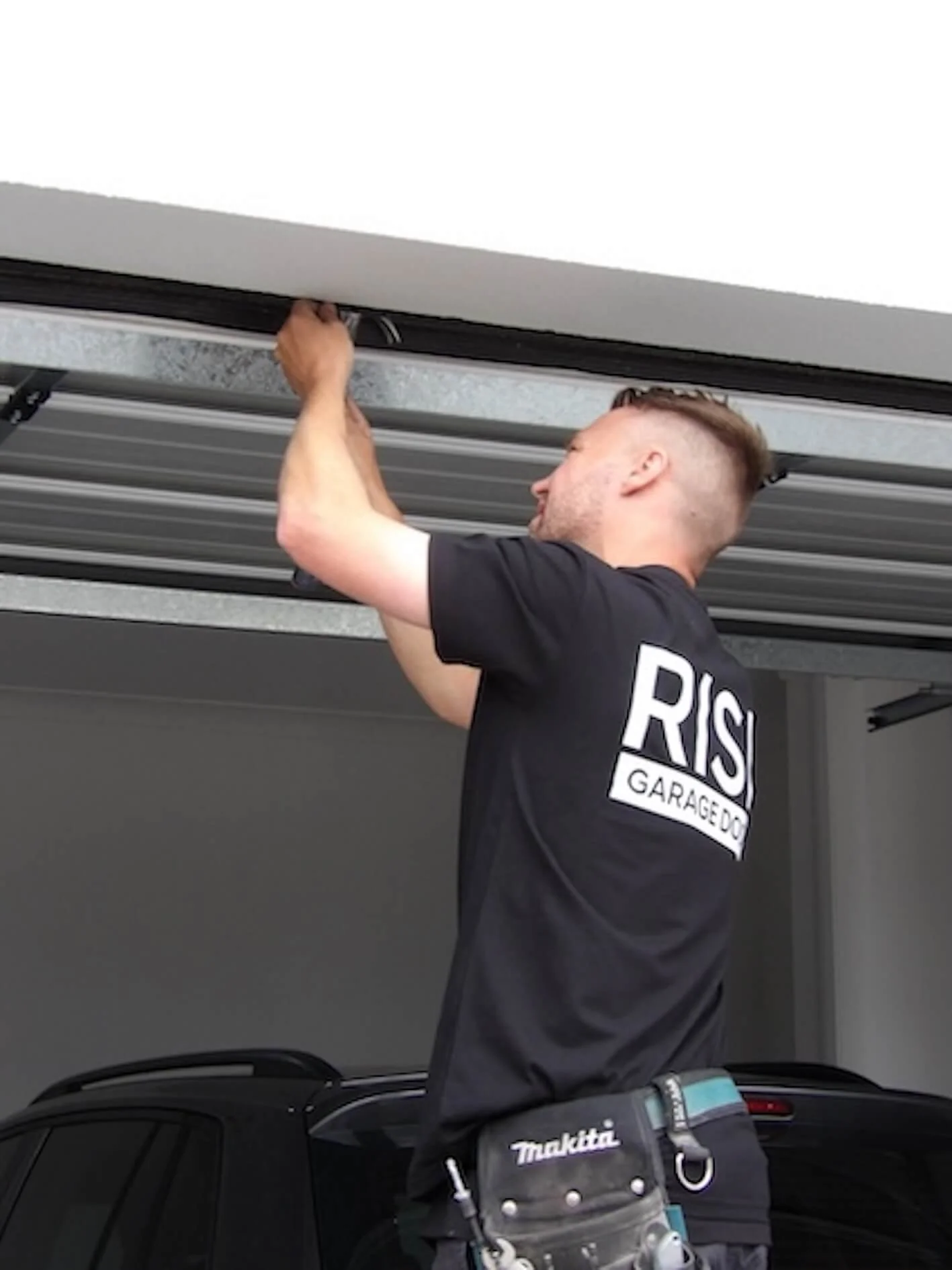 A man working on a garage door, wearing a black shirt with 'RIS GARAGEDO' on the back, and a tool belt with a Makita tool attached.