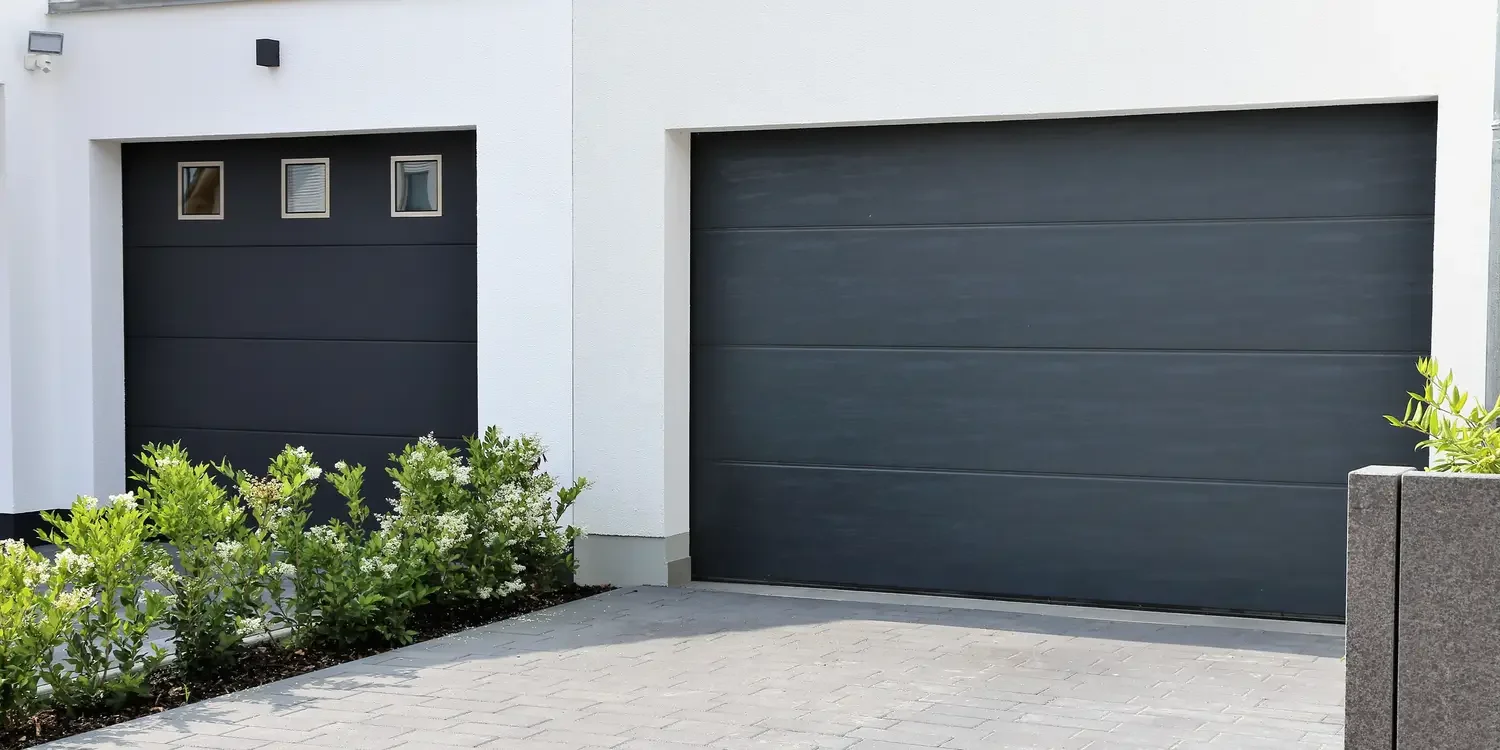Modern white house with two large black garage doors and small windows, surrounded by green plants and shrubs.